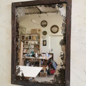 May include: A rectangular mirror with a distressed, dark brown wooden frame and a chipped, aged glass surface. The mirror reflects a workshop with tools and other objects.