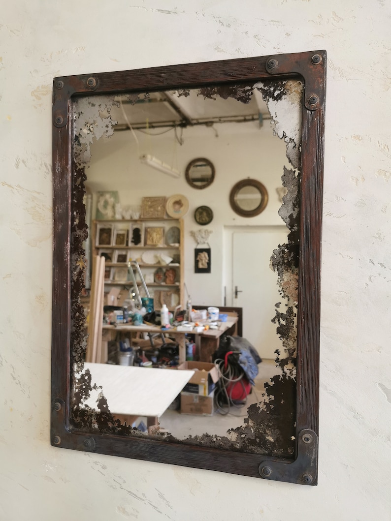 May include: A rectangular mirror with a distressed, dark brown wooden frame and a chipped, aged glass surface. The mirror reflects a workshop with tools and other objects.