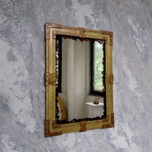 May include: A gold-toned mirror with a decorative, ornate frame. The frame is made of metal and has a hammered texture. The mirror is rectangular and hangs on a textured wall.