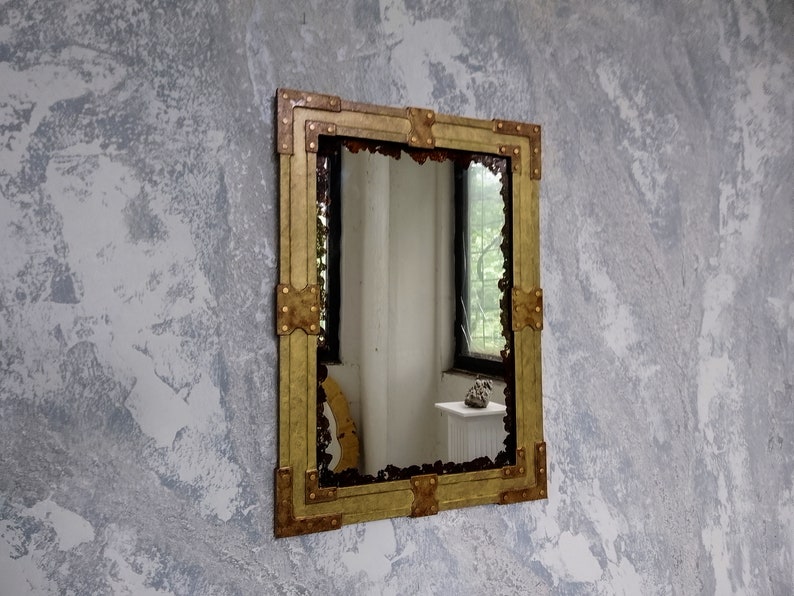 May include: A gold-toned mirror with a decorative, ornate frame. The frame is made of metal and has a hammered texture. The mirror is rectangular and hangs on a textured wall.