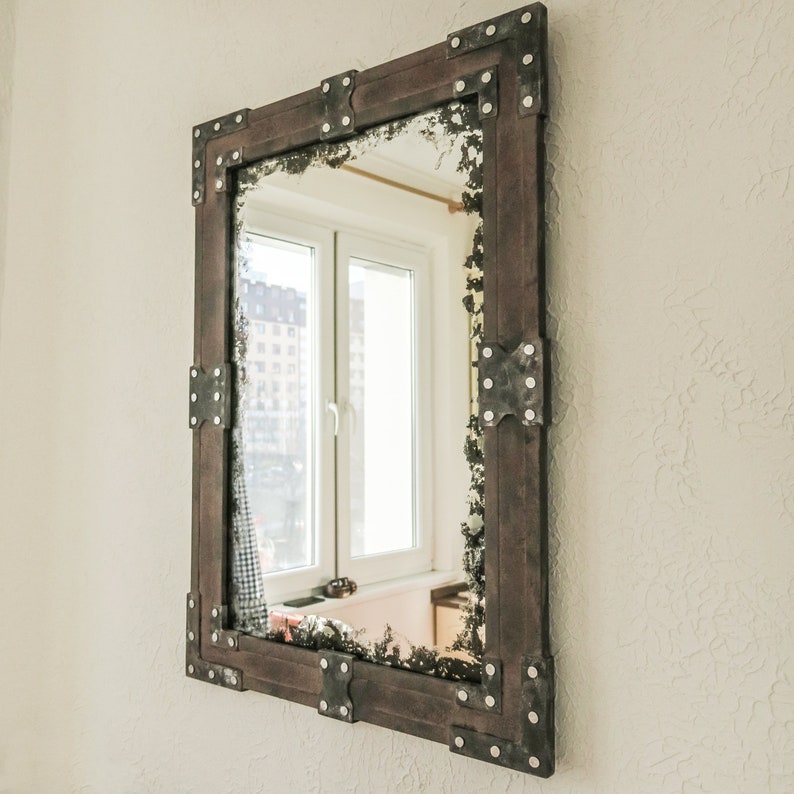 May include: A rectangular mirror with a dark brown wooden frame and metal accents. The frame has decorative metal pieces in each corner. The mirror reflects a window with white trim.