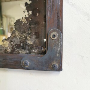 May include: A rustic mirror with a distressed, aged look. The mirror is framed in dark wood with metal corner brackets and screws. The mirror has a chipped and cracked surface, giving it a unique and vintage aesthetic.