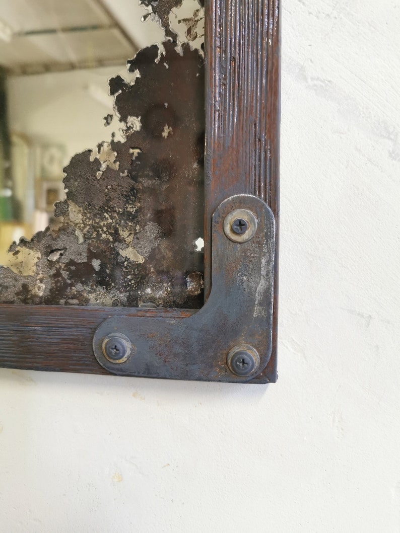 May include: A rustic mirror with a distressed, aged look. The mirror is framed in dark wood with metal corner brackets and screws. The mirror has a chipped and cracked surface, giving it a unique and vintage aesthetic.