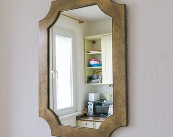 May include: A gold-toned, ornate mirror with a beveled edge and a scalloped frame. The mirror reflects a kitchen with white cabinets, a window, and appliances.