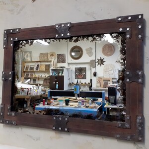 May include: A rustic wooden mirror with a distressed, antique-style finish. The frame is adorned with metal corner brackets and rivets. The mirror reflects a workshop with tools and equipment.