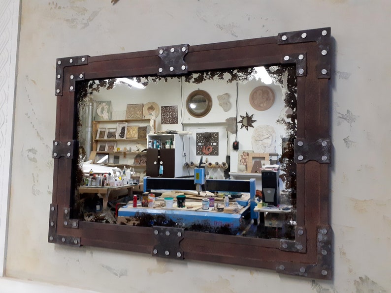 May include: A rustic wooden mirror with a distressed, antique-style finish. The frame is adorned with metal corner brackets and rivets. The mirror reflects a workshop with tools and equipment.