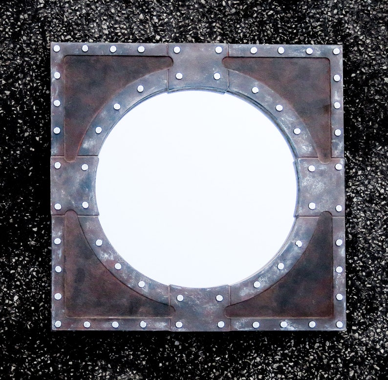 May include: A round mirror with a rustic, industrial-style frame. The frame is made of metal and has a distressed, aged look. The mirror is surrounded by a series of rivets.