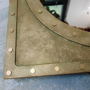 May include: A gold-toned metal mirror with a rounded top edge and a distressed finish. The mirror is framed with a thick metal border and has decorative rivets around the edge.