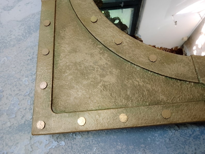 May include: A gold-toned metal mirror with a rounded top edge and a distressed finish. The mirror is framed with a thick metal border and has decorative rivets around the edge.