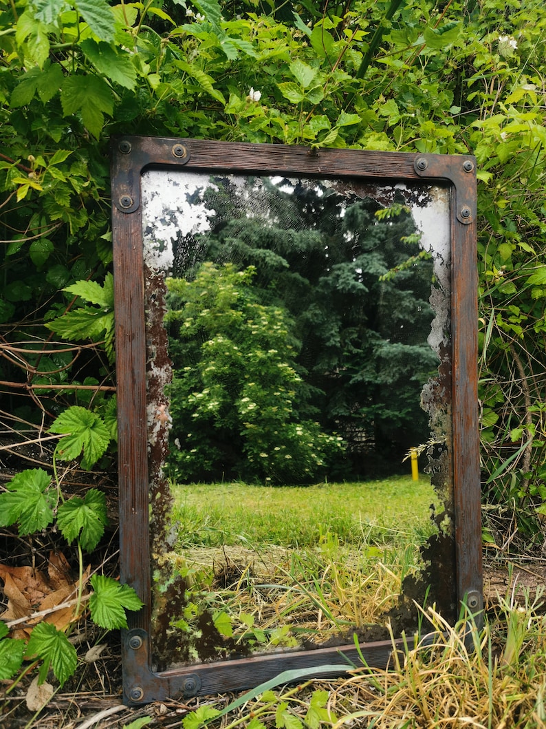 May include: A rustic wooden framed mirror with a weathered, aged look. The mirror reflects a green grassy area with trees in the background.
