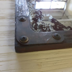 May include: A close-up of a vintage mirror with a distressed, aged, silver-toned frame. The frame has a wood-like texture and is secured with three visible screws. The mirror is slightly cracked and shows signs of wear and tear.