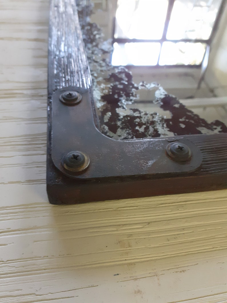 May include: A close-up of a vintage mirror with a distressed, aged, silver-toned frame. The frame has a wood-like texture and is secured with three visible screws. The mirror is slightly cracked and shows signs of wear and tear.