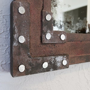 May include: A rectangular mirror with a distressed, rusty metal frame. The frame has a layered design with rivets along the edges. The mirror has a slightly aged, cloudy appearance.