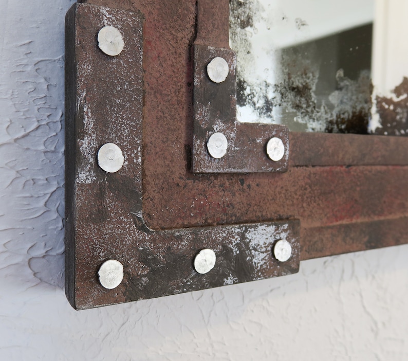 May include: A rectangular mirror with a distressed, rusty metal frame. The frame has a layered design with rivets along the edges. The mirror has a slightly aged, cloudy appearance.