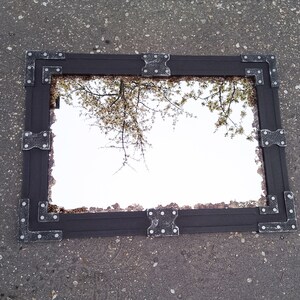 May include: A black framed mirror with ornate metal accents. The mirror reflects a view of a tree with white blossoms.