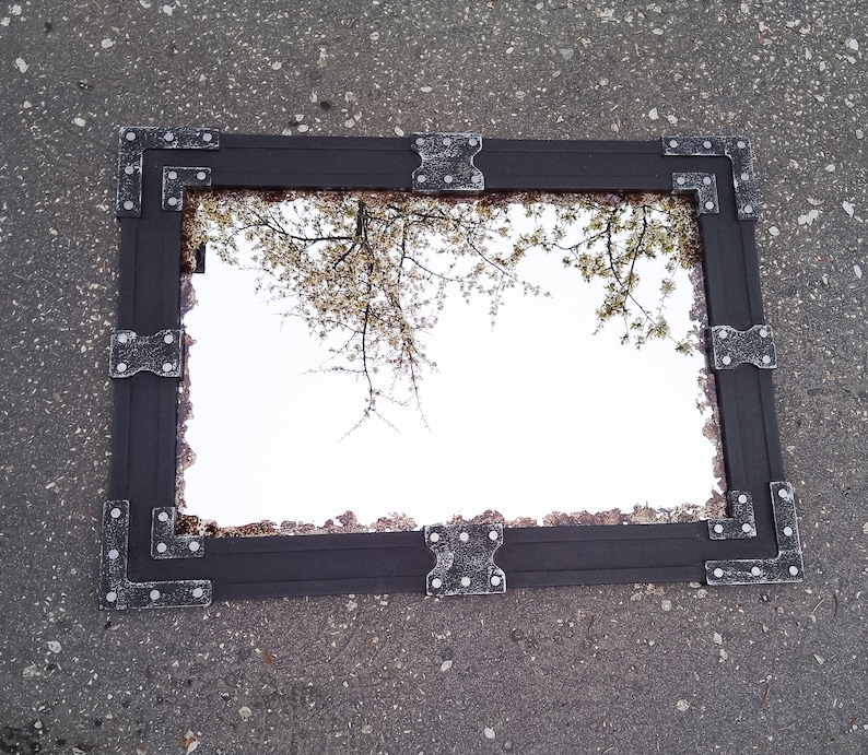 May include: A black framed mirror with ornate metal accents. The mirror reflects a view of a tree with white blossoms.