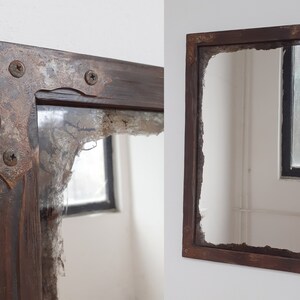 May include: A rustic wooden mirror with a distressed, chipped, and cracked glass surface. The frame is dark brown with metal corner accents. The mirror is hanging on a white wall.