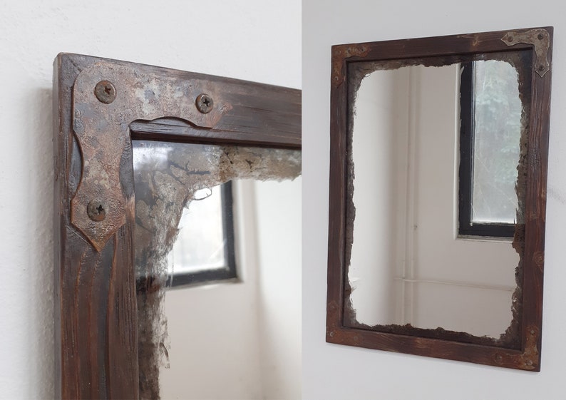 May include: A rustic wooden mirror with a distressed, chipped, and cracked glass surface. The frame is dark brown with metal corner accents. The mirror is hanging on a white wall.