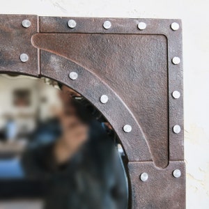 May include: A round mirror with a rustic, aged metal frame. The frame is decorated with silver rivets and has a slightly rounded edge. The mirror is reflecting a blurry image of a person.