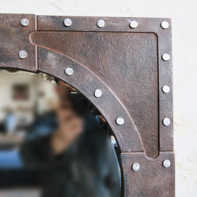 May include: A round mirror with a rustic, aged metal frame. The frame is decorated with silver rivets and has a slightly rounded edge. The mirror is reflecting a blurry image of a person.