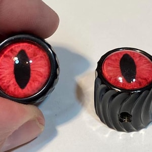 May include: Two black decorative knobs with a red eye design. One knob is held by a hand. The eye design features a red iris with a black oval pupil. The knobs have a black, textured, cylindrical base.