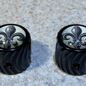 May include: Two black, decorative knobs with a fleur-de-lis design. The knobs have a glossy finish and a spiral carved pattern. The fleur-de-lis is black and white, set against a light background.