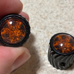 May include: Two black guitar knobs with a raised, textured design. The knobs have a clear, round top with an orange and black pentagram design.