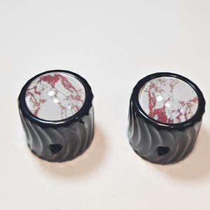 May include: Two black, cylindrical bottle stoppers with a swirled design. Each stopper has a clear, circular top with a red and white marbled pattern. The stoppers are displayed on a white surface.