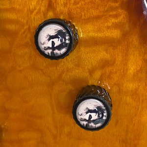 May include: Two black guitar knobs with a white and black abstract design, set against a warm, amber-colored wood grain background. The knobs have a textured, ridged edge.