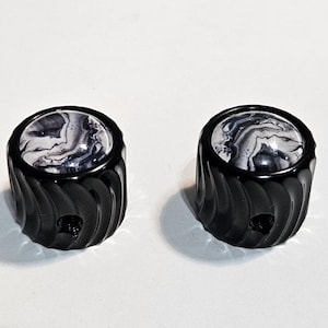 May include: Two cylindrical control knobs featuring a matte black exterior with a textured, spiraled grooved pattern. The top surface of each knob is inset with a smooth, circular dome displaying a swirling black, white, and gray marble pattern.