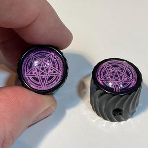 May include: Two black guitar knobs with a pink pentagram design. The knobs have a textured, ridged surface.