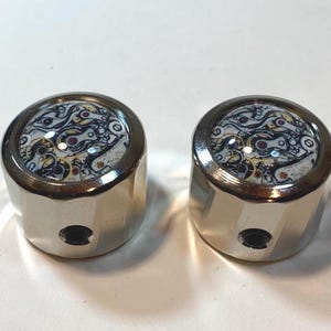 May include: Two silver-toned knobs with a watch mechanism design on the top. The knobs have a cylindrical shape with a small hole on the side. The watch mechanism design features a mix of black, white, and gold colors.