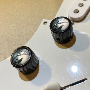 May include: Two black guitar knobs with a white background. The knobs have a silver ring around the edge and a graphic of a crow and a cross in a forest setting.