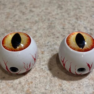 May include: Two white, spherical decorative objects with a detailed eye design. The eyes feature a yellow iris with a black pupil, surrounded by red and orange detailing. Red vein-like patterns are painted on the white surface.