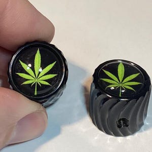 May include: Two black metal knobs with a green marijuana leaf design. The knobs have a textured surface.
