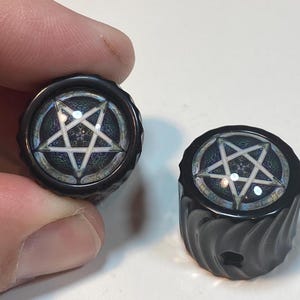 May include: Two black guitar knobs with a white pentagram design. The knobs have a textured surface and a black background.