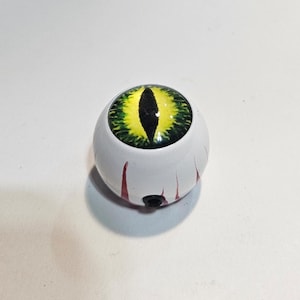 May include: A spherical object resembling a human eyeball with a white base and red vein-like markings. The center features a prominent iris with a yellow and green gradient and a vertical black pupil. A small black circular port is visible at the base.