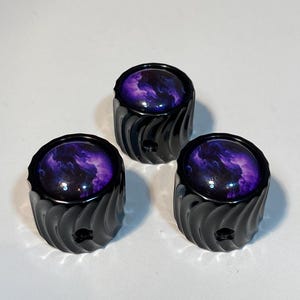 May include: Three black knobs with a swirling, textured design. Each knob features a circular, glossy top with a purple and white nebula pattern. The knobs are likely for a piece of furniture or equipment.