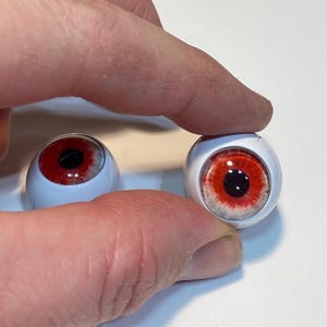 May include: Two doll eyes with a white sclera, a red iris, and a black pupil. The eyes are being held by a person's fingers. The eyes are a craft supply.