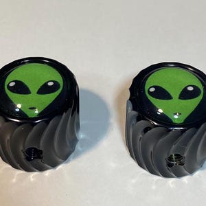 May include: Two black, decorative knobs with a green alien face design. The alien has large black eyes and a small mouth. The knobs have a spiral design on the sides and a threaded hole in the center.