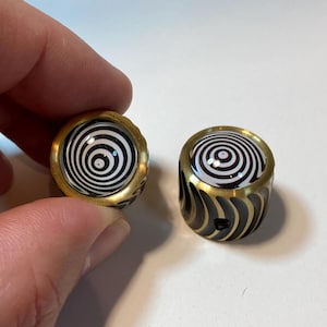 May include: Two brass beads with a black and white spiral design. The beads have a gold-colored rim and a black, curved pattern on the sides. One bead is held by a hand.