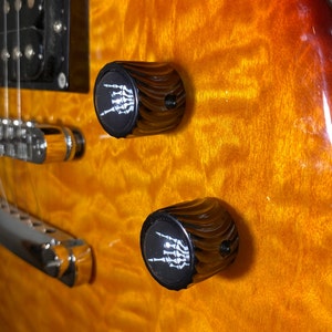 May include: Close-up of two black guitar knobs with a white skeleton hand design. The knobs are on a brown and orange guitar.