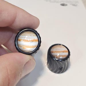 May include: Two cylindrical control knobs featuring a glossy, circular inlay depicting the planet Jupiter with its characteristic orange, white, and brown banded cloud patterns. The knobs have a black, ribbed, and textured outer metal casing.