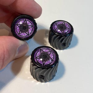 May include: Three black bottle stoppers with a purple and black pentagram design. The stoppers have a textured, ridged body and a circular top with the graphic. One stopper is held by a hand.