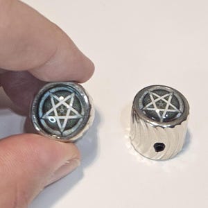 May include: Two cylindrical silver-toned metal knobs featuring a circular inlay on the top. Each inlay displays a white five-pointed star pentagram set against a dark, textured background. The sides of the knobs have a vertical, wavy ribbed pattern.