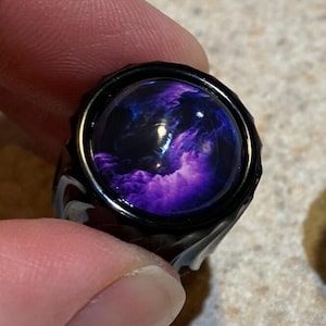 May include: A black ring with a round, domed center featuring a swirling purple and blue design. The ring's band has a textured, marbled appearance. The ring is held between fingers.
