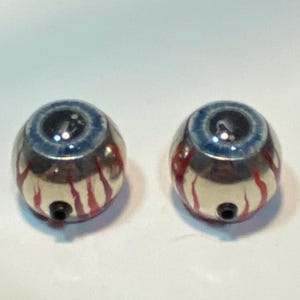 May include: Two round, metallic eye-shaped objects with blue irises and black pupils. Red vein-like patterns are painted on the surface. Each has a small black hole at the bottom, suggesting a possible attachment point.