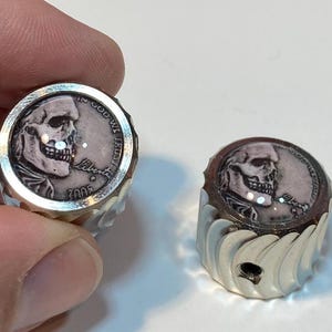 May include: Two silver-colored metal knobs with a skull design on the top. The skull is a profile view of a human skull with a detailed design. The knobs have a textured, ridged design on the sides. The text "In God We Trust" and "Liberty" are visible on the top knob.