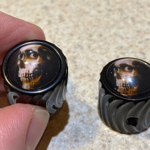 May include: Two black guitar knobs with a skull design. The knobs have a textured, wavy pattern.