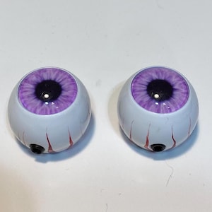 May include: Two white, spherical eyeballs with purple irises and black pupils. Red veins are painted on the white surface. Each eyeball has a small black hole at the bottom.